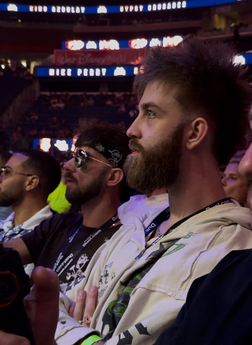 A bearded man with spiky hair, wearing a hoodie and lanyard, intently watches an event at a stadium alongside others, including a friend in sunglasses and a bandana. The "Disrael" text visible in the background ties into the post's playful request of "Can I get a GM?" suggesting the viewer greet the focused spectator.