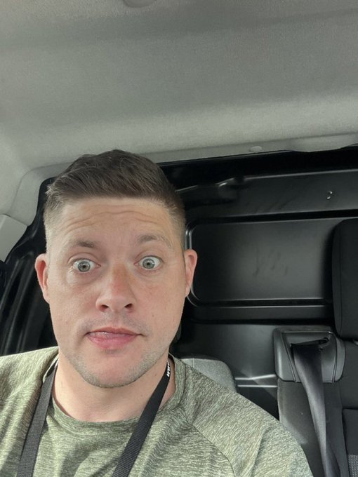 A man with short brown hair and wide eyes taking a selfie inside a vehicle, wearing an olive green shirt with a lanyard around his neck.