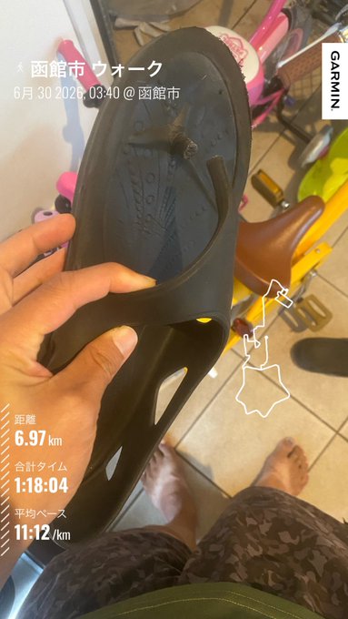 A hand holds up a worn black sandal/shoe, with a Japanese workout stats overlay showing 函館市 ウォーク, 6.97 km, 1:18:04 time, and 11:12/km pace via GARMIN.