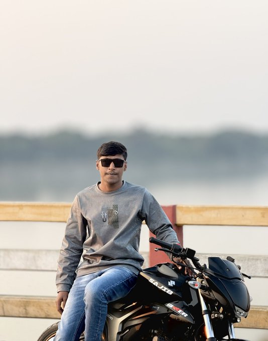 A young man wearing sunglasses and a gray sweatshirt sits on a black Suzuki motorcycle with a hazy landscape in the background.