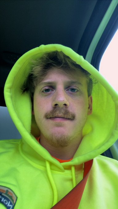 A close-up selfie shows a man with a mustache wearing a bright neon green hoodie, sitting inside a vehicle.