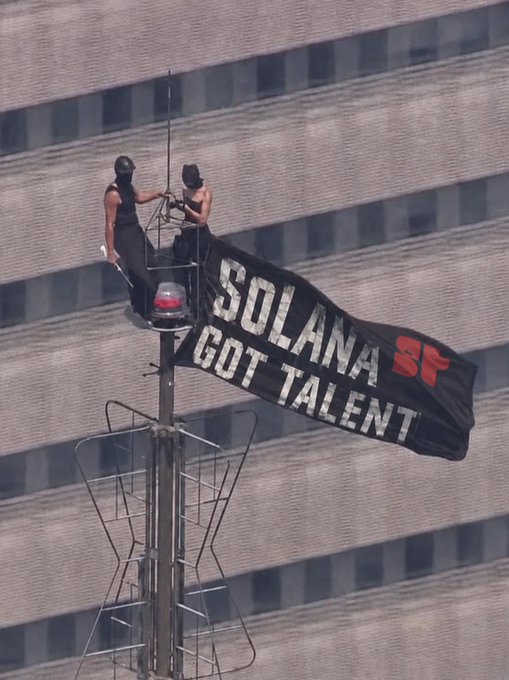 Two people stand atop a tall antenna or tower, unfurling a large black banner that reads "SOLANA SF GOT TALENT" with a red "SF" logo, performing a daring promotional stunt to advertise the Solana Got Talent event.