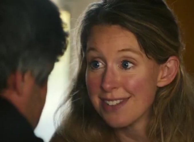 A close-up of a smiling woman with blue eyes looking at someone whose shoulder and head are visible in the foreground.