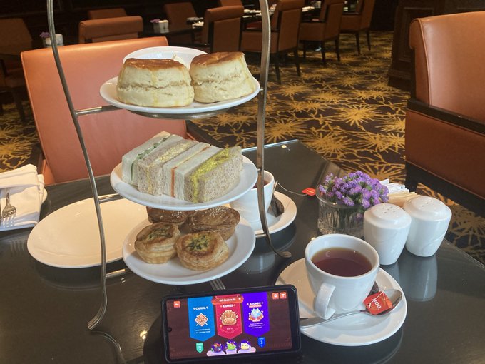 A traditional afternoon tea setup featuring a three-tiered stand with scones, finger sandwiches, and pastries, accompanied by a teacup on a table in what appears to be a lounge, with a smartphone displaying a sports/team app in the foreground.