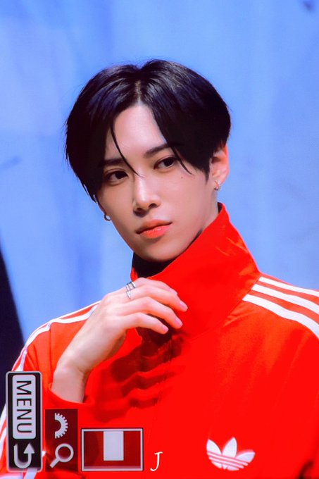 A young man with dark hair, wearing a red Adidas track jacket with white stripes, poses with his hand near his chin against a blue backdrop, with "MENU," "J," and camera icons visible at the bottom of the frame. The hashtags indicate this image is connected to ALD1 (Alpha Drive One) member ARNO, likely captured during a fan event or photoshoot.