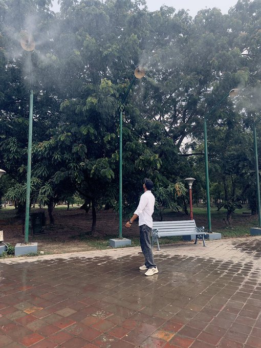 A man in a white shirt stands in a park plaza paved with red tiles, surrounded by green trees and mist from sprinklers under a cloudy sky.