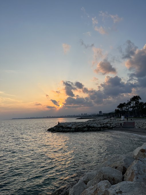 Sunset photo over a coastal city, showing the sun breaking through clouds over a bay with a stone breakwater, palm trees, and distant buildings on the horizon.