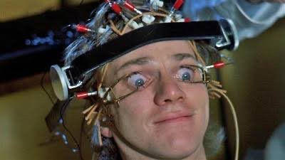 A comedic close-up of a man wearing a head apparatus with multiple wires and electrodes attached to his head and face, displaying an exaggerated expression.