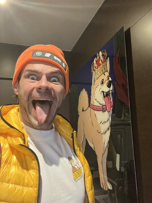 A man in a yellow puffer vest and orange beanie sticks his tongue out next to a poster of a crowned corgi dog.