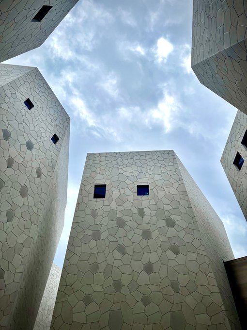 A low-angle view looking up at modern architectural structures with white, Voronoi-patterned facades, irregular dark windows, and pointed tops against a cloudy sky.
