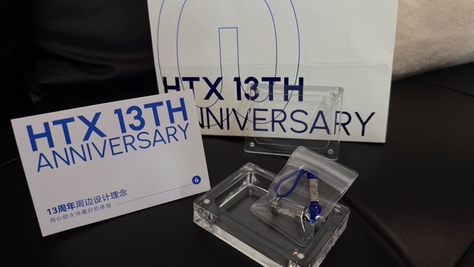 An 'HTX 13TH ANNIVERSARY' display features promotional materials with Chinese text alongside a small clear acrylic display case containing a blue beaded accessory.