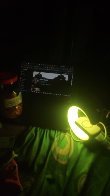 In a dark room, a laptop screen displays a YouTube video, illuminated by the green glow of a ring light resting on a green fabric surface.