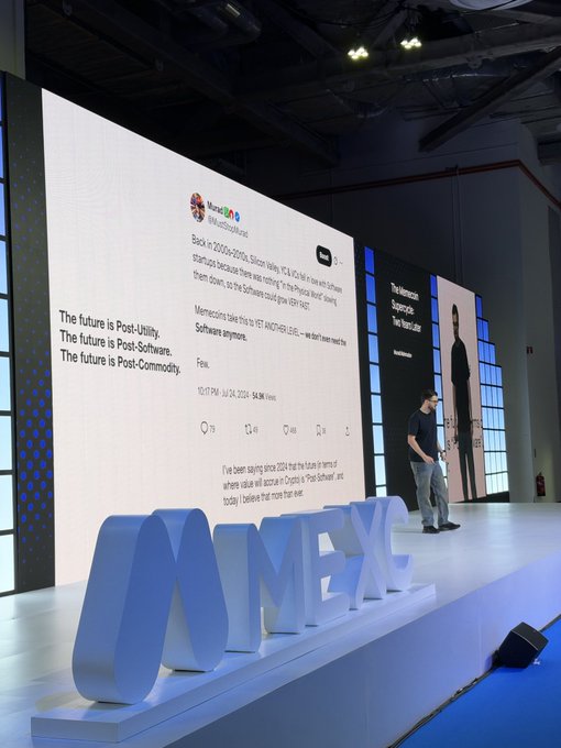 A speaker stands on stage at a tech conference in front of a giant screen displaying a tweet from @MustStopMurad about "MMXX" (the "NMEX" letters are visible in the foreground as decor), with text emphasizing "The future is Post-Utility, The future is Post-Software, The future is Post-Commodity" — perfectly matching the caption of him "teeing up" another bold prediction.