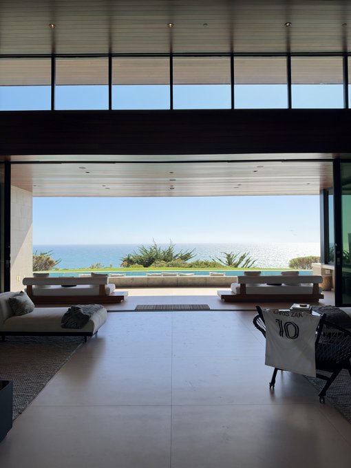 An open-plan luxury living room looks out through a large window to an infinity pool overlooking the ocean, with a football jersey bearing "GYOKU ZAK 10" draped over a chair.