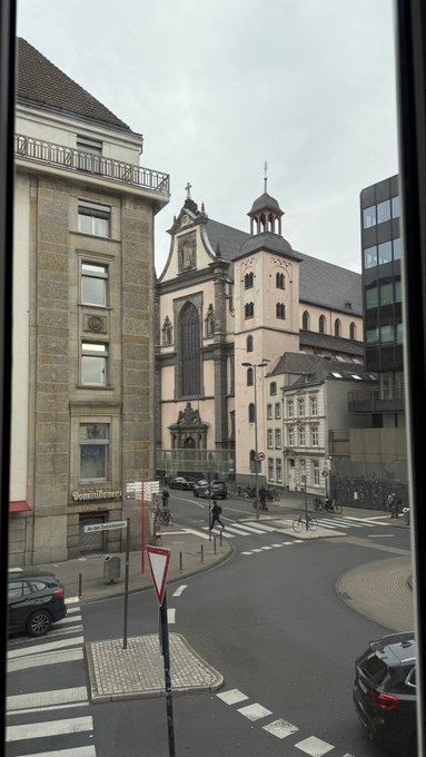 A view from a window of a European street corner featuring historic stone buildings, a Gothic church with an ornate tower, parked cars, and pedestrian crossings on an overcast day.