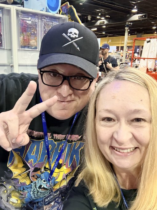 A selfie of a man wearing a black skull cap and glasses flashing a peace sign next to a smiling woman with long blonde hair, taken at what appears to be a comic convention.