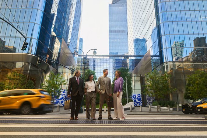 Caption: A low-angle shot captures four people in business attire standing together on a city street crosswalk, surrounded by tall glass skyscrapers, with a yellow taxi blurred in motion on the left side.