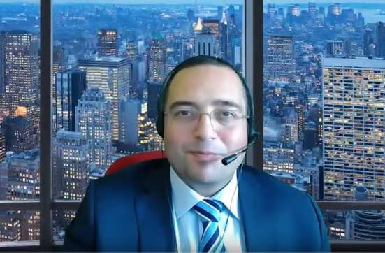 A man wearing glasses, a headset, and a suit with a striped tie appears on a video call or livestream, with a backdrop of a city skyline at dusk. The image relates to the Turkish post announcing a live broadcast scheduled for 16:00 Turkey time before the US stock market opening, covering topics like $btc, $SOL, and gold, with the man likely being the host preparing to address these topics.