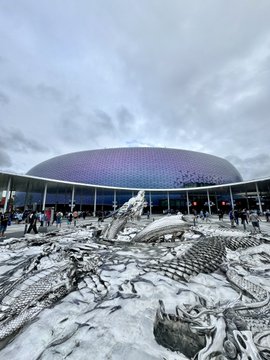 A large modern stadium with a colorful facade serves as the backdrop for an elaborate monochrome sculpture featuring a dragon in the foreground, where OKX branding is prominently displayed across the venue and surrounding areas during what appears to be a Manchester City promotional event.