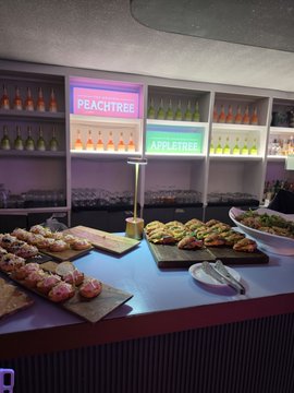 A catering or buffet display featuring rows of appetizers like crostini and bruschetta, with neon signs in the background reading 'PEACHTREE' and 'APPLETREE'.
