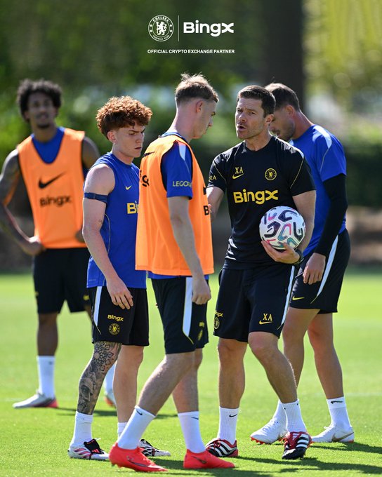 A coach in a black Chelsea FC training kit labeled "BingX" holds a colorful soccer ball while speaking with players in orange and blue training gear, with the "BingX OFFICIAL CRYPTO EXCHANGE PARTNER" banner and Chelsea crest displayed at the top of the image.