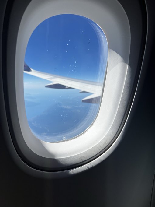 A view through an oval airplane window showing the wing of the aircraft against a bright blue sky with a layer of clouds below.