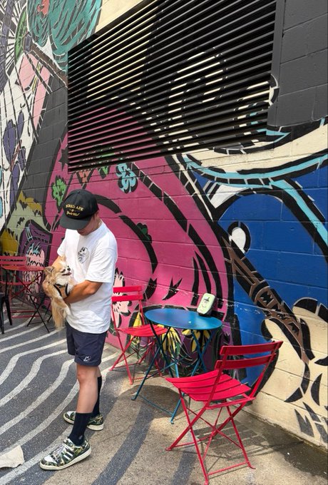 A person in a black cap holding a small dog stands next to a blue table and red chairs outside a building covered in vibrant street art murals.