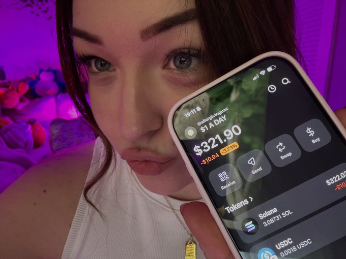 A woman with blue eyes holds up a smartphone displaying a crypto trading app showing $321.90, with the username "@allegedzoggg" and tokens including Solana and USDC visible.