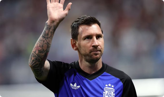 Lionel Messi in a purple Adidas AFA jersey waves with his tattooed right arm in what appears to be a stadium setting.