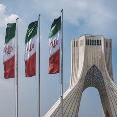 Three Iranian flags wave in the foreground against the backdrop of Tehran's iconic Azadi Tower, symbolizing the nation at the center of the report about Iran's increasing reliance on cryptocurrency, particularly USDT and bitcoin, to navigate cross-border transactions amid tightening U.S. sanctions.