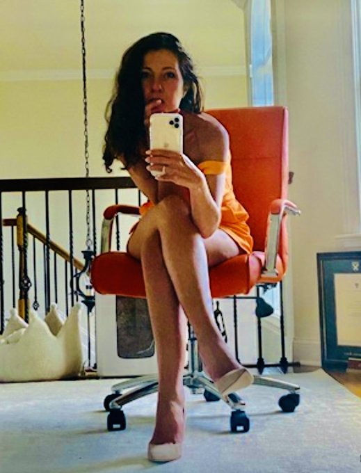 A woman in an orange dress sits in an orange chair taking a mirror selfie with a white iPhone in a home interior setting.