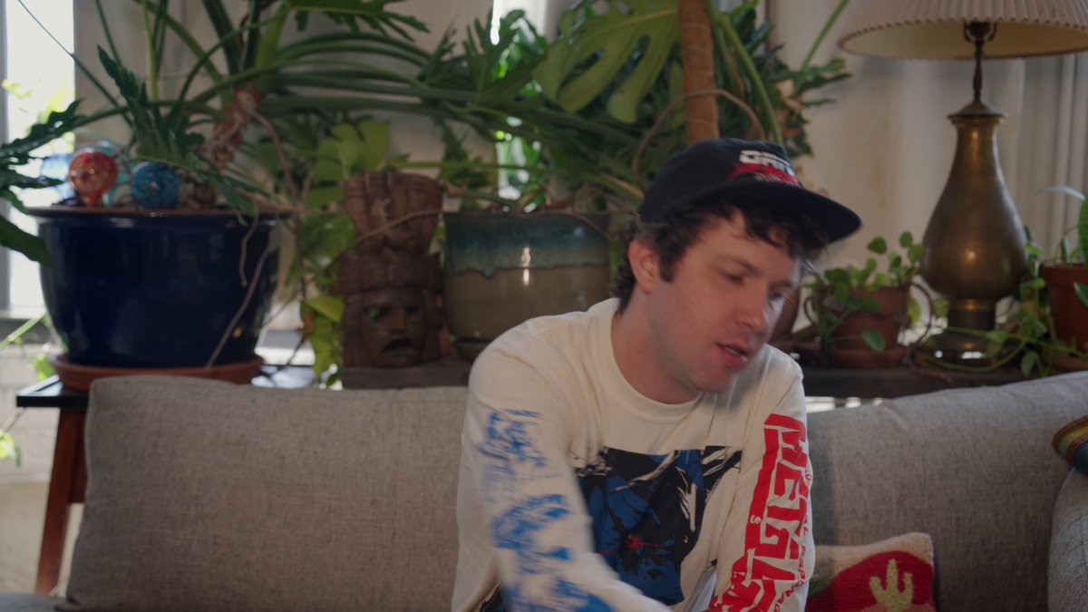 In this image, there is a young man wearing a baseball cap sitting comfortably on a couch with his hands resting behind him. The man appears to be enjoying a moment of relaxation at home. Next to the couch, there are two potted plants adding a touch of greenery and life to the scene. On the right side of the image, there is an additional potted plant and a lamp providing soft lighting in the room.