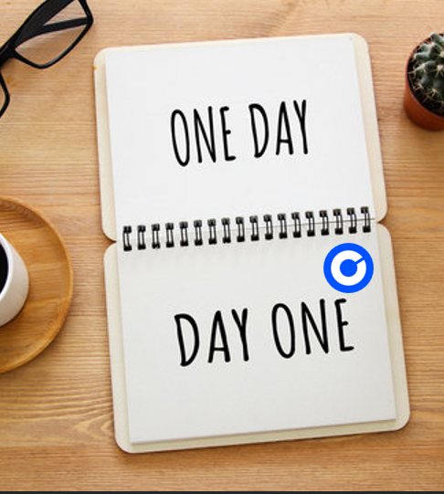 An open spiral notebook on a wooden desk with the text 'ONE DAY' on the top page and 'DAY ONE' on the bottom page, accompanied by glasses, a cup of coffee, and a small cactus.