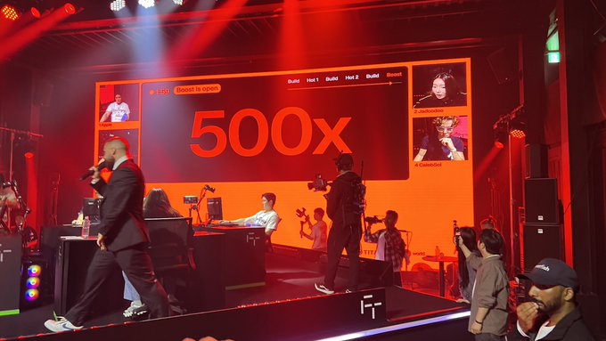 An esports tournament stage is bathed in red lighting, featuring a large screen displaying '500x' along with player names 'Hoti', 'Build', and 'Rooted5'.