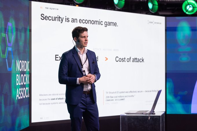 A man in a suit giving a presentation at the Nordic Blockchain Association, with a slide reading 'Security is an economic game' and showing 'Cost of attack' economics.