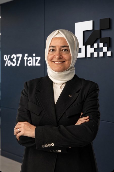 A portrait of a smiling woman wearing a white hijab and a black suit jacket, standing with arms crossed in front of a dark blue wall displaying the text '%37 faiz'.