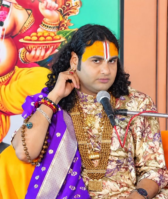 A man dressed in ornate traditional attire with devotional markings, jewelry, and a purple shawl speaks into a microphone at a public event. The image accompanies a post about donating a 30,000-crore fixed deposit and Bitcoin profits to Sanatan Dharma and Maharaji; no readable text or numbers appear in the image.