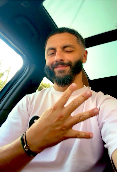A bearded man in a white t-shirt sits inside a car, smiling with his eyes closed while holding up his right hand in a casual gesture.