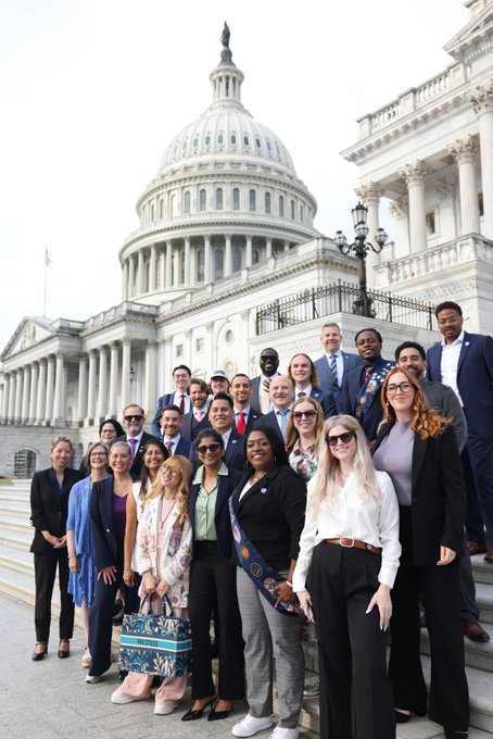 A group photo of around 20 people in professional attire posing on the steps of the U.S. Capitol building in front of its iconic white dome.