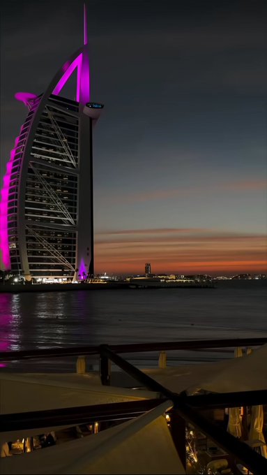 The Burj Al Arab hotel in Dubai illuminated in pink and purple lights against a sunset sky, viewed from across the water.