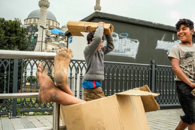 Two barefoot street children play joyfully with cardboard boxes near a mosque in the background, their carefree laughter embodying the post's message to embrace the day with simple, unfiltered happiness.