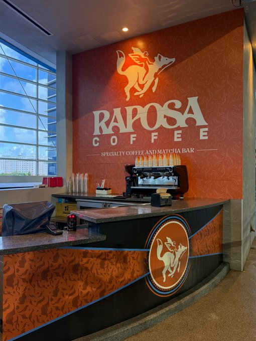 The interior of a Raposa Coffee specialty coffee and matcha bar, featuring an orange wall with the brand's dog-and-rider logo, an espresso machine, and large windows.