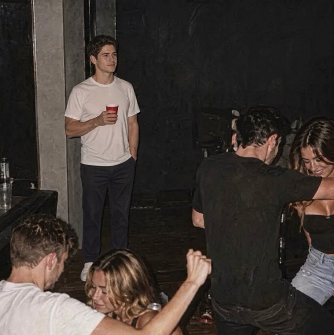 A young man in a white t-shirt and dark pants holds a red cup while standing against a wall at a social gathering.