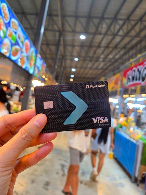 A hand holds up a black "Bitget Wallet" Visa Platinum card with a blue chevron design in a vibrant food market, with menu boards and food stalls blurred in the background. The image visually reinforces the caption's message by capturing the card in a real-world setting where everyday purchases—like satisfying food cravings—become seamless transactions.