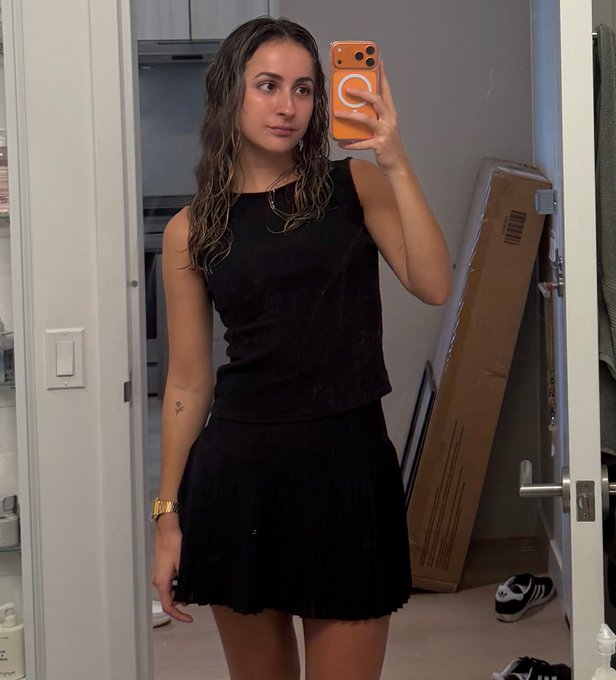 The image features a woman taking a selfie with her cell phone, holding it up to take a picture of herself in the mirror. She appears to be wearing a black dress or skirt and has a short ponytail. The background shows a door leading into another room, suggesting that she is indoors. There are also several shoes visible near the doorway, indicating that this might be her home or apartment.