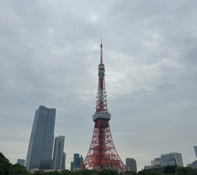 A view of Tokyo Tower, the iconic red and white lattice communications tower, set against an overcast sky with modern skyscrapers and greenery visible at its base.
