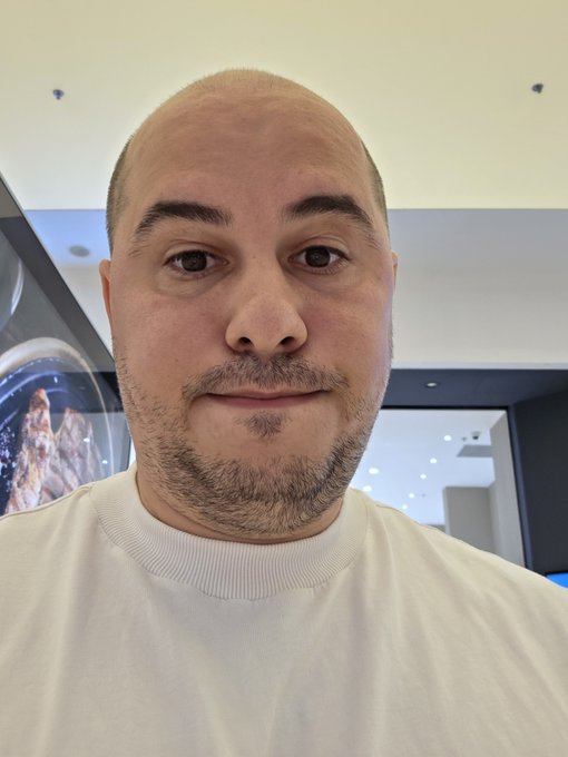 A close-up selfie of a bald man with a goatee wearing a white shirt, photographed indoors with circular light fixtures visible in the background.