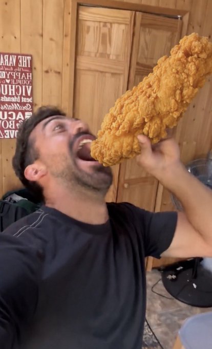 A man with dark hair eating a very large piece of fried chicken, holding it up to his mouth in what appears to be a kitchen with wooden cabinets.