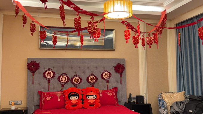 A bedroom decorated for a Chinese wedding, featuring red hanging banners with the '囍' (double happiness) character, red bedding, and a headboard adorned with Chinese characters meaning 'we are engaged'.