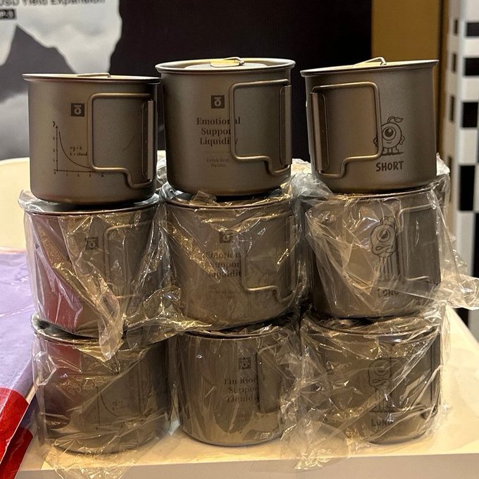A stack of metallic camping-style cups labeled "Emotional Support Liquidity," "SHORT," and "LONG" with a math graph design, arranged in plastic-wrapped piles at what appears to be a promotional booth. The image relates to the post's mention of StandX and BSC (Binance Smart Chain) recovery, likely symbolizing the "emotional support" StandX provides during market volatility through its SIP 5B program and customizable Vault visibility features.